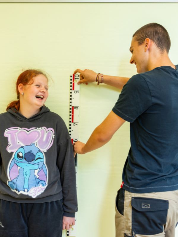 Leerling wordt lachend opgemeten door begeleider in een lesruimte op school.