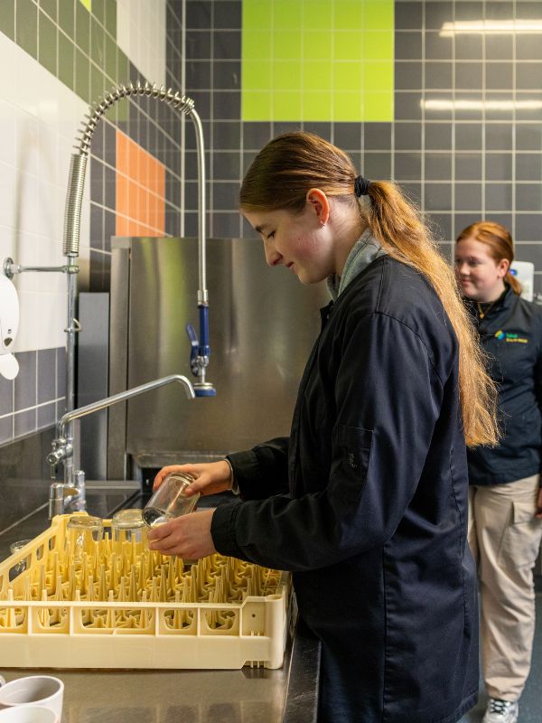Praktijkles in de spoelkeuken Leerlingen werken samen in de spoelkeuken en plaatsen glazen in een rek tijdens een praktijkles horeca of facilitair werk