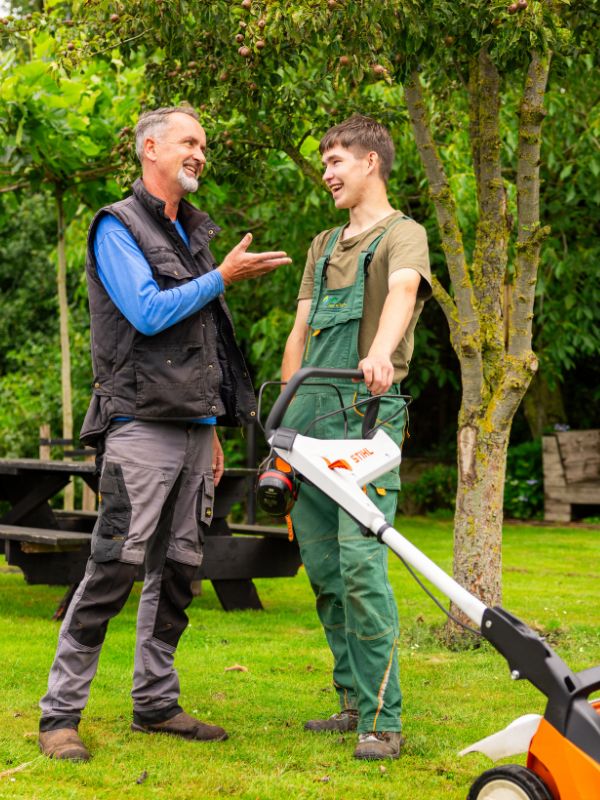 Samen werken in het groen Leerling in werkkleding staat met een grasmaaier in een groene tuin en praat met een begeleider tijdens een praktijkmoment buiten